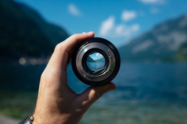 person holding a camera lens framing a view of mountains and a body of water