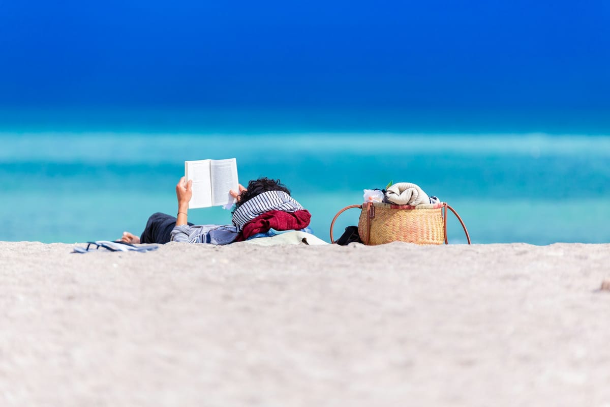 person lying on the beach reading a book against the turquoise blue of the ocean