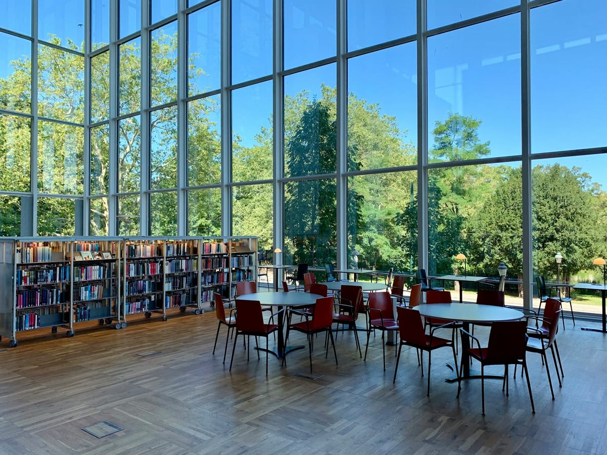 bookshelves and chairs and tables in a glass-walled room looking out at green trees and a bright blue sky