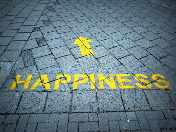 Happiness written in yellow capital letters and topped by a yellow arrow on a grey brick sidewalk