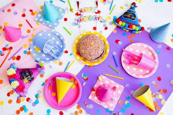 colourful assortment of paper party ware, cone hats, cake, and a happy birthday banner on a confetti-filled surface