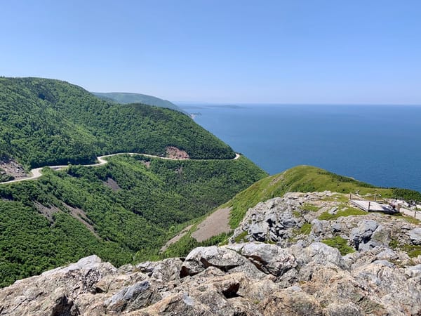 A view of the Atlantic Ocean beyond large mountains with a winding road through them from atop the Skyline Trail in Cape Breton