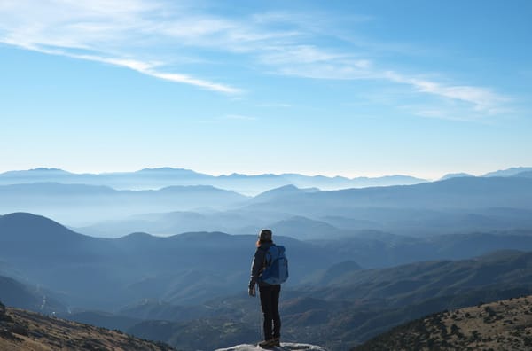 person on a mountaintop looking out at a silhouette of mountain ranges in various shades of blue under a blue sky