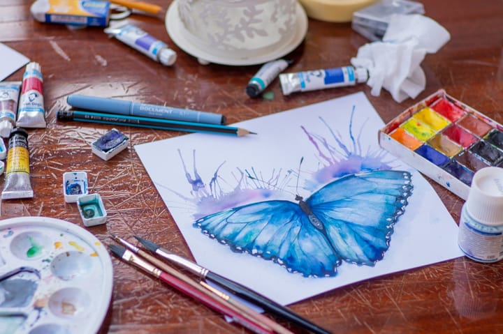 watercolour painting of a blue butterfly on a table and surrounded by art supplies